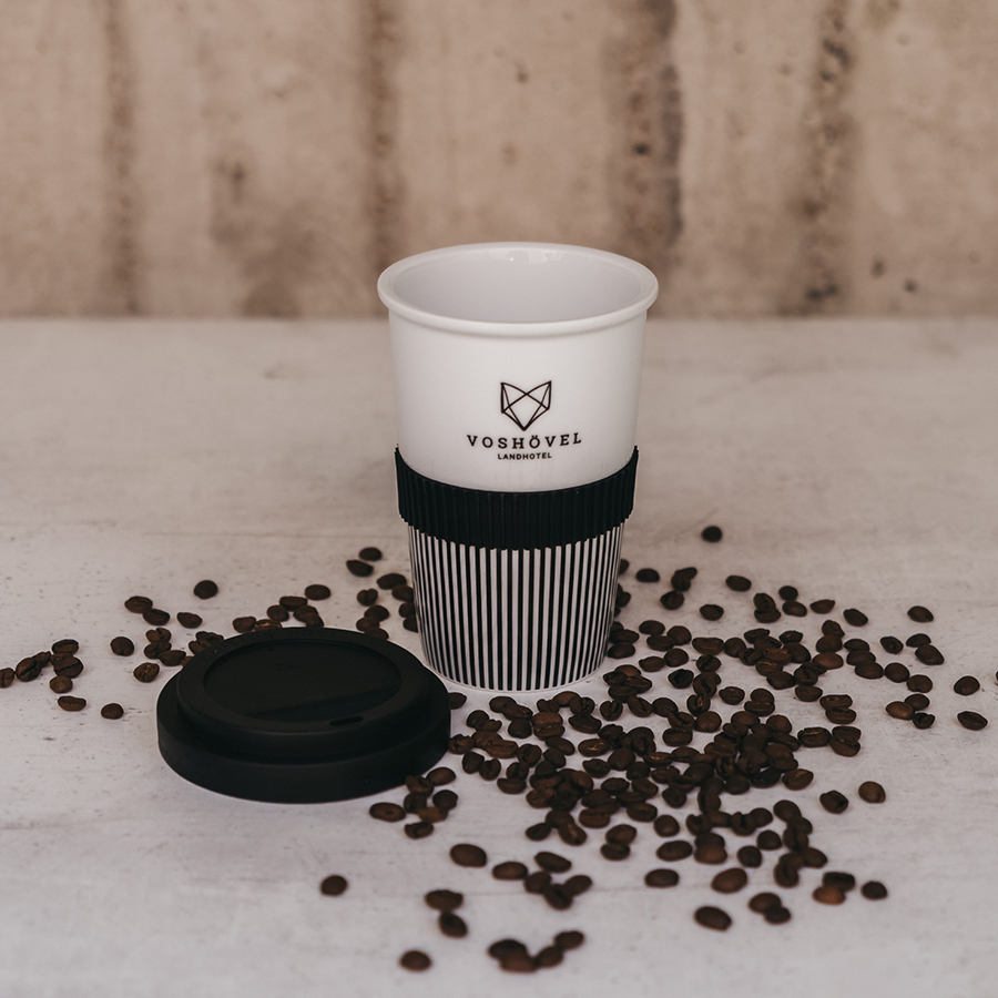 Schwarz-weißer Kaffe-to-go Becher mit dem Voshövel Logo und geöffnetem Deckel in Kaffeebohnen